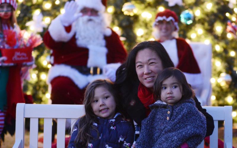 Laura Mead, middle, sits with her daughters Angelina, 5, left, and Samantha, 2, on the right, as Santa waves in the background. Due to COVID-19 restrictions and adjustments, children were not able to sit on Santa’s lap and tell him what they wanted this year. Instead, they were allowed to sit on a bench in front of him and Mrs. Claus for photo opportunities.
