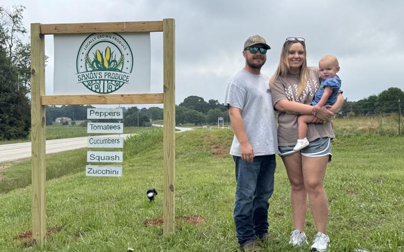 The Saxon family (L-R) Garrett Saxon, Breanna Saxon and Annalee Saxon stand next to the Saxon’s Produce sign located at 8701 Bowman Highway in Dewy Rose. Saxon’s Produce first opened on Memorial Day 2025 and sells double-yolk eggs, pickles and produce. (Photo/Adriana Cascio)