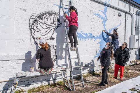 Nora Winkler, from left, Alondra Arevalo, on the ladder, and Katie Eidson, right paint a mural on the rear exterior wall of Little Japan on Depot Street in downtown Hartwell recently as restaurant owners Treena and Tommy Nguyen look on. 