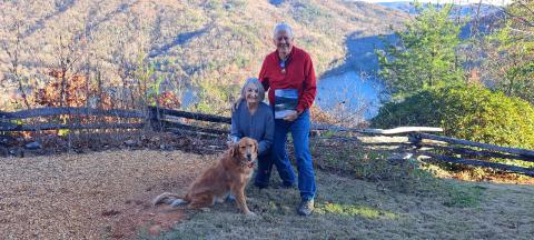 A picture viewing a snowy mountain range and snow-covered fence at Lake Burton taken by Chip Goen of Clayton is now featured on the cover of the Winter 2024 Lake Living magazine. Lin Goen submitted the photograph to the contest. The Goens are pictured here with their dog Charley, the Lake Living magazine cover and $100, with the same backdrop in a different season.