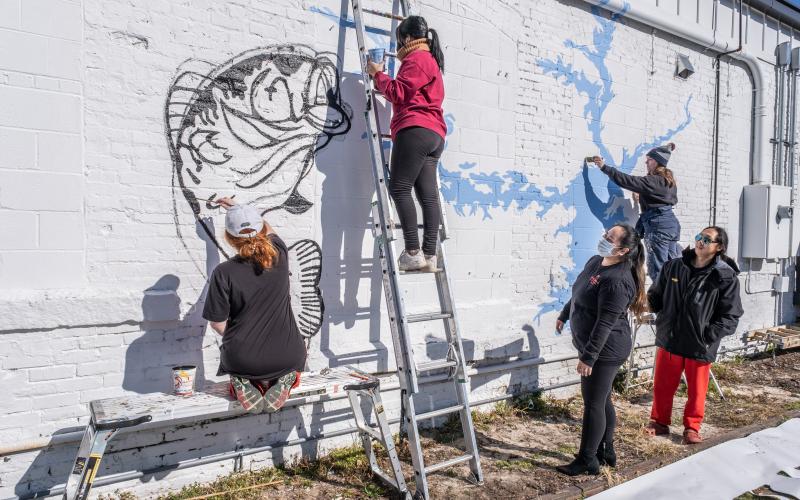 Nora Winkler, from left, Alondra Arevalo, on the ladder, and Katie Eidson, right paint a mural on the rear exterior wall of Little Japan on Depot Street in downtown Hartwell recently as restaurant owners Treena and Tommy Nguyen look on. 