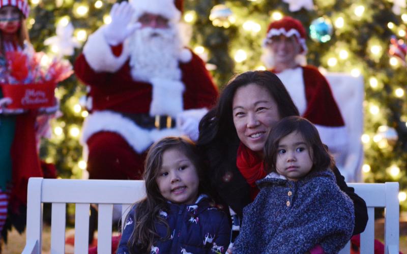  Laura Mead, middle, sits with her daughters Angelina, 5, left, and Samantha, 2, on the right, as Santa waves in the background. Due to COVID-19 restrictions and adjustments, children were not able to sit on Santa’s lap and tell him what they wanted this year. Instead, they were allowed to sit on a bench in front of him and Mrs. Claus for photo opportunities. 